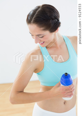 Fit smiling woman holding water bottle at fitness studio 9124553