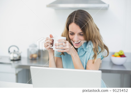 Amused beautiful woman using her notebook while holding a cup 9125452