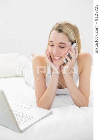 Smiling blonde woman lying on her bed phoning using her notebook 9125725