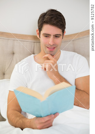 Relaxed young man reading book in bed 9125782