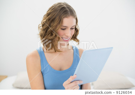 Cheerful cute woman using her blue tablet while sitting on her b 9126034