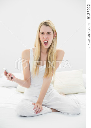 Shocked blonde woman holding her smartphone while sitting on her 9127022