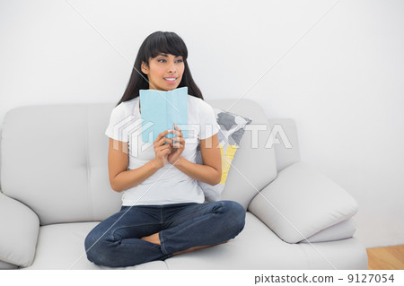 Thoughtful young woman holding a book sitting on couch 9127054