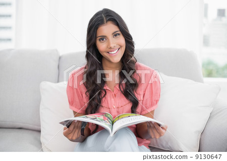 Smiling cute brunette sitting on couch holding magazine Smiling cute brunette sitting on couch holding magazine 9130647