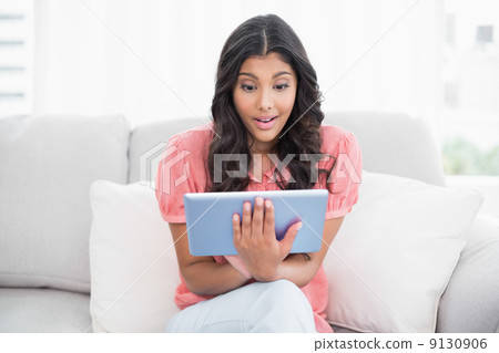 Excited cute brunette sitting on couch using tablet 9130906