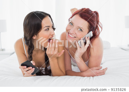 Shocked woman looking at a cheerful friend while on call in bed Shocked woman looking at a cheerful friend while on call in bed 9135826