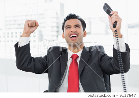 Businessman cheering with telephone receiver at office Businessman cheering with telephone receiver at office 9137647