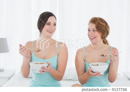 Smiling female friends with salad bowls sitting on bed 9137786