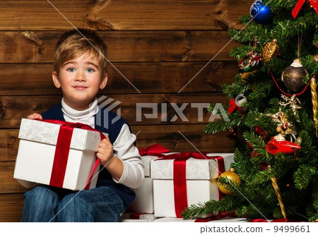 Little boy with gift box under christmas tree in wooden house interior Little boy with gift box under christmas tree in wooden house interior 9499661