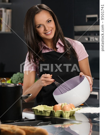 Young woman in apron whisking eggs for a dough on a modern kitchen 9501741