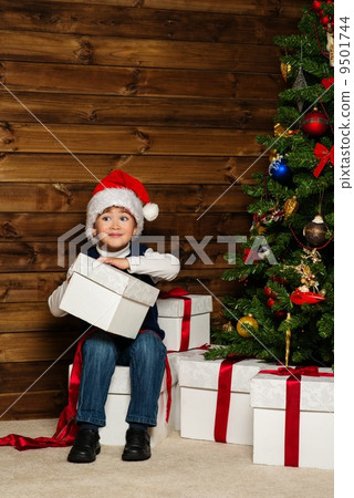LIttle boy in Santa hat with gift box under christmas tree in wooden house interior 9501744