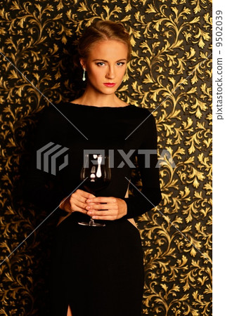 Beautiful young girl with glass of wine wearing black evening dress in luxury interior 9502039