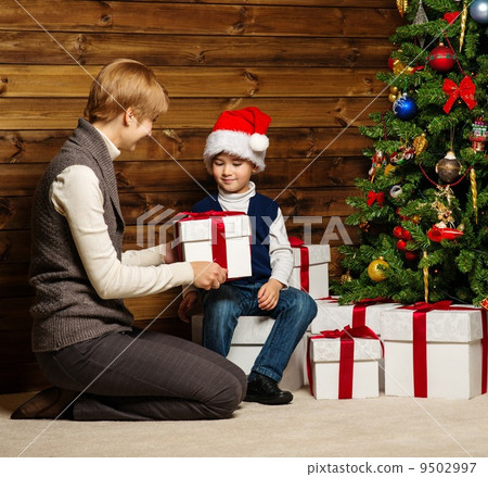Mother and her lIttle boy in Santa hat with gift box under christmas tree in wooden house interior 9502997
