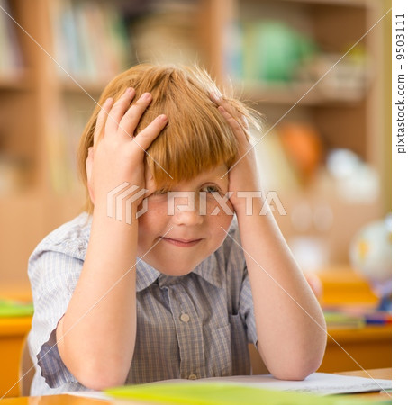 Little redhead schoolboy behind school desk during lesson Little redhead schoolboy behind school desk during lesson 9503111