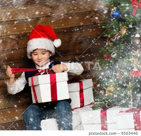Little boy in Santa hat opening gift box under christmas tree in wooden house interior 9503440