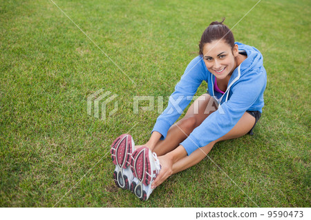 Pretty sporty woman stretching her legs while sitting on the gra 9590473