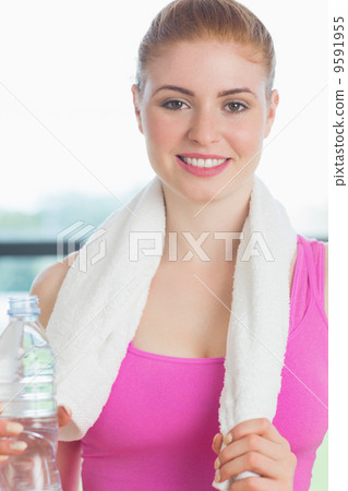 Woman with towel around neck holding water bottle in fitness stu 9591955