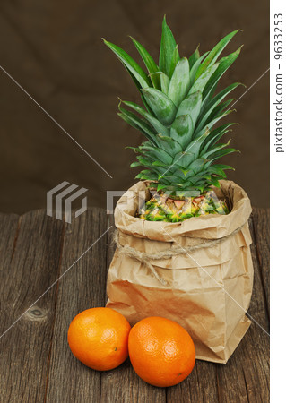 Ripe pineapple in paper bag with oranges on wooden table. 9633253