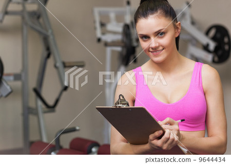 Smiling female trainer writing on clipboard in gym 9644434