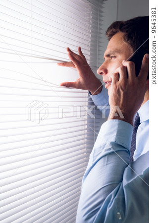 Businessman peeking through blinds while on call in office Businessman peeking through blinds while on call in office 9645381