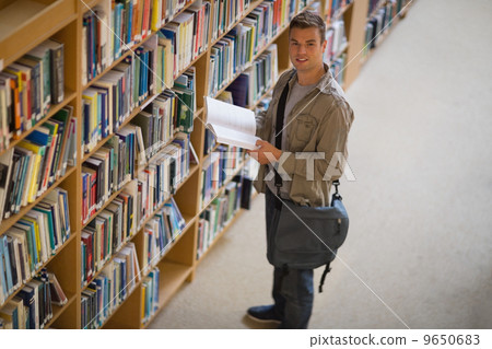 Student holding a book from shelf in library smiling at camera 9650683