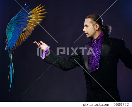 Young handsome brunette magician man in stage costume with his trained parrot Young handsome brunette magician man in stage costume with his trained parrot 9689722