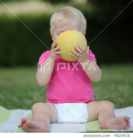 Adorable little girl is sitting on a carpet in a park and biting on a half of melon 9820928