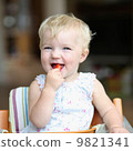 Funny happy healthy toddler sitting in high feeding chair biting on cherry tomato 9821341