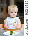 Cute baby girl sitting in a high feeding chair biting on delicious freshly cooked broccoli  9821510