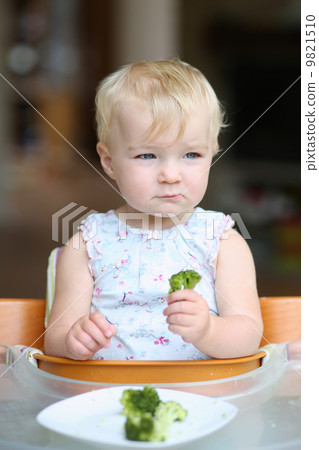 Cute baby girl sitting in a high feeding chair biting on delicious freshly cooked broccoli Cute baby girl sitting in a high feeding chair biting on delicious freshly cooked broccoli 9821510