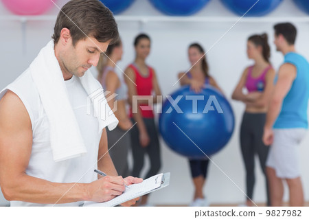 Trainer writing in clipboard with fitness class in background 9827982