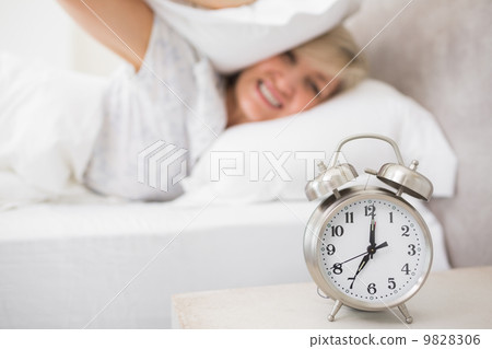 Woman covering ears with pillow with alarm clock in foreground 9828306