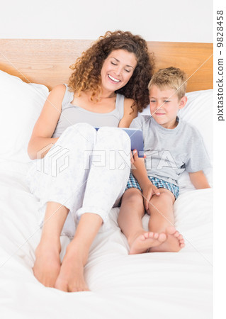 Happy mother and son sitting on bed with tablet pc 9828458