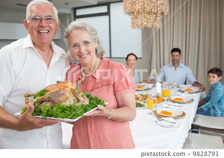 Grandparents holding chicken roast with family at dining table Grandparents holding chicken roast with family at dining table 9828791
