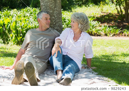 Mature couple sitting together against tree at park 9829243