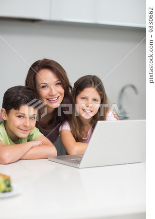 Smiling mother with kids using laptop in kitchen 9830662