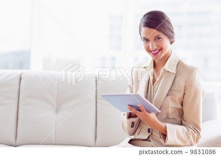 Stylish businesswoman sitting on sofa using tablet smiling at ca Stylish businesswoman sitting on sofa using tablet smiling at ca 9831086