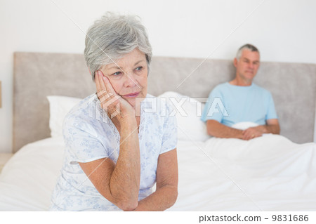 Sad senior woman on bed with husband in background Sad senior woman on bed with husband in background 9831686