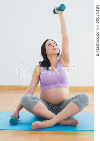 Pregnant brunette sitting on exercise mat lifting hand weight Pregnant brunette sitting on exercise mat lifting hand weight 10025193