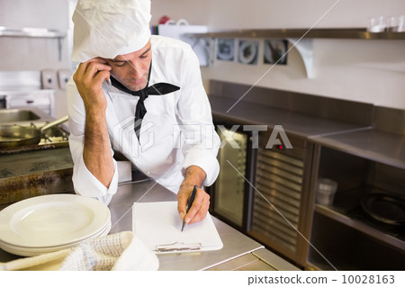 Cook writing on clipboard while using cellphone in kitchen Cook writing on clipboard while using cellphone in kitchen 10028163