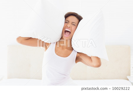 Screaming brunette sitting on bed covering ears with pillow Screaming brunette sitting on bed covering ears with pillow 10029222