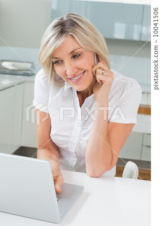 Casual woman using laptop while on call in kitchen 10041506