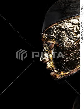 Styled Woman's Face Covered Golden Foil over Black Background. Mystery 10077622
