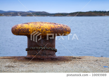 Old rusted yellow mooring bollard with chain on concrete pier 10093989