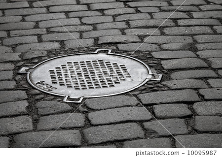 Old sewer manhole on dark cobblestone pavement 10095987