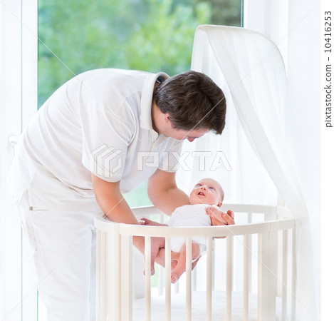 Happy young father putting his smiling baby in a white round cri Happy young father putting his smiling baby in a white round cri 10416253