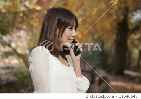 A woman standing at a park that has autumn leaves and hanging up a cell phone 11046645