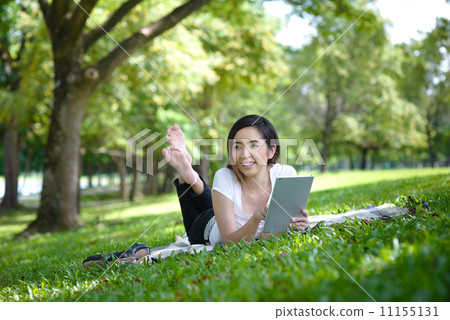 Young woman in a park wearing casual laying on a mat using mobile device tablet iPad 11155131
