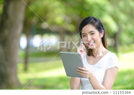 Young woman in a park wearing casual sitting on a mat using mobile device tablet iPad 11155164