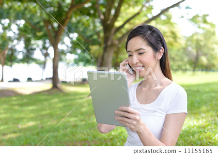 Young woman in a park wearing casual sitting on a mat using mobile device tablet iPad 11155165